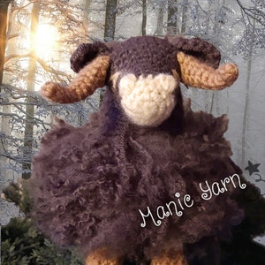 May include: A brown crocheted sheep with a white face and brown horns. The sheep is sitting on a green surface with a snowy forest background. The text "Manic Yarn" is visible on the sheep's body.
