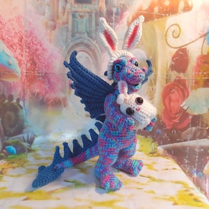 May include: A crocheted blue and pink dragon wearing a white bunny hat and holding a white bunny in its arms. The dragon has wings and a long tail.