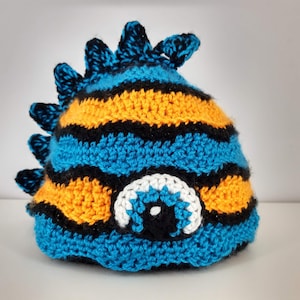 May include: A blue, black, and orange crocheted hat with a large white eye and black pupil. The hat has a spiky design on top.