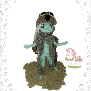 May include: A crocheted amigurumi doll with a brown and gray hooded coat, light blue skin, and white hair. The doll is standing on a brown surface. The text "Manic Yarn" is visible in the image.