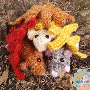 May include: A collection of handmade crochet mice in various colors, including brown, gray, and cream, surrounded by crochet leaves in red, yellow, and orange. The mice have black eyes and pink noses and ears. The image is set against a natural, earthy background.