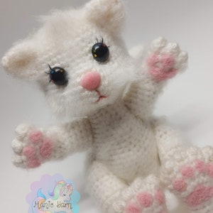May include: White crocheted cat amigurumi toy with pink paw pads and black eyes. The toy is sitting on a white surface.