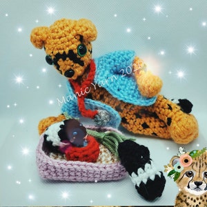 May include: Crocheted cheetah figurine with orange and black spots, wearing a blue jacket and red scarf. A small crocheted basket holds a baby cheetah. The text "Manic Yarn 2020" is visible.