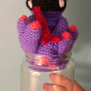May include: Purple crocheted monster with a red tongue sticking out, sitting inside a clear glass jar.