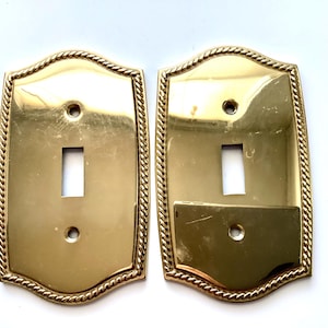 May include: Two brass-colored light switch covers. Each cover has a rectangular opening for the switch and a decorative rope-like border. The covers have a shiny, reflective surface and are mounted on a white background.