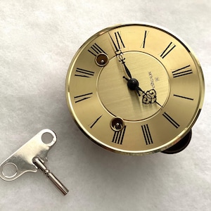 May include: A round gold-toned clock face with black Roman numerals and ornate black hands. The dial features the text Montreux and two winding holes. A separate silver-toned metal winding key with a T-shaped handle is positioned to the left.