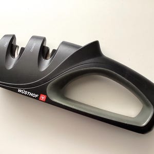 May include: A black and gray Wüsthof knife sharpener with a handle. The sharpener has two slots labeled "FINE" and "COARSE". The handle is gray and the Wüsthof logo is red.