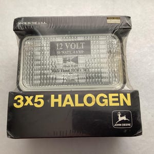 May include: A rectangular 3x5 halogen work light in black packaging. The clear lens features text reading 12 VOLT, 50 WATT, 4 AMP, and Wide Flood. The black box displays the John Deere logo, the text 3X5 HALOGEN, and Made in the U.S.A. in white.