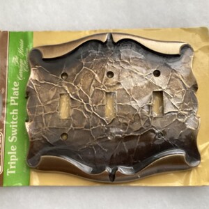May include: A bronze-toned triple switch plate with a textured, cracked finish, mounted on its original cardboard packaging. The packaging features the Amerock brand logo, the text Triple Switch Plate, and the Carriage House Collection.