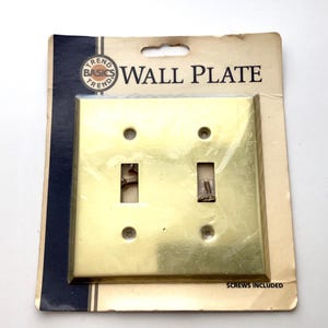 May include: A brass-colored double light switch wall plate with screws included, mounted on a cardboard backing. The packaging reads "TREND BASICS" and "WALL PLATE".