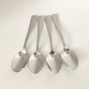 May include: Four stainless steel spoons with a reflective, polished finish. Each spoon has a ribbed design on the handle and a rounded bowl. The spoons are arranged on a white surface.