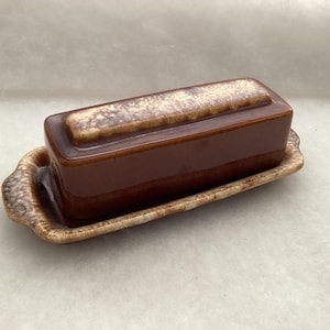 May include: A rectangular ceramic butter dish featuring a solid dark brown base and a matching lid. The lid and the edges of the tray display a mottled, textured cream and brown glaze pattern, providing a contrast to the smooth, dark brown sides.