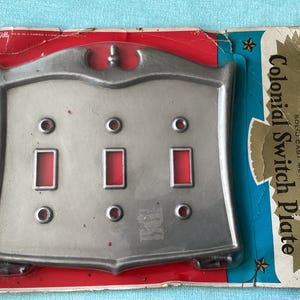 May include: A silver-colored Colonial Switch Plate made of solid cast metal. The plate has three rectangular openings for switches and is mounted on a red, white, and blue cardboard backing.