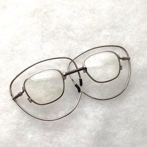 May include: Silver-toned metal eyeglasses featuring a unique double-frame design. The inner frames hold clear lenses, while the larger outer frames are connected by a central spring-loaded bridge. Small black nose pads are attached to the inner frame.