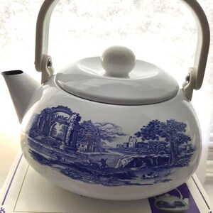 May include: A white enamel teapot featuring a blue transferware-style landscape print depicting ruins, trees, and figures. The teapot has a rounded body, a matching lid with a spherical knob, and a white arched handle attached to the sides.