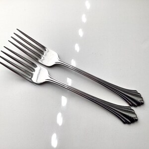 May include: Two stainless steel forks with a decorative handle design. The forks have four tines and are angled towards the upper right. The handles have a scalloped design at the base. The forks are on a white surface.