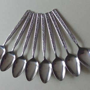 May include: A collection of eight stainless steel spoons arranged in a fan shape. Each spoon has a polished, oval bowl and a handle with an ornate, engraved design. The spoons are displayed on a light-colored surface.
