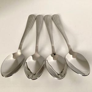 May include: Four shiny, silver spoons with elongated oval bowls and ridged handles. The spoons are arranged in a row, reflecting light. A set of elegant flatware for dining.