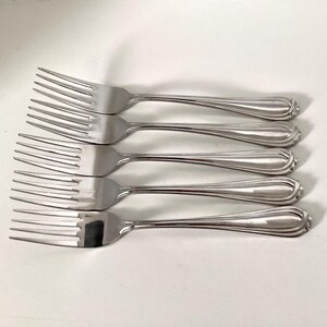 May include: Five stainless steel forks arranged in a row. Each fork has four tines and a handle with a decorative, ridged design. The forks are made of shiny, polished metal, reflecting the light.