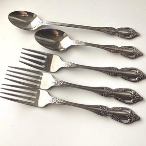 May include: Five pieces of silverware, including two spoons and three forks, are arranged on a white surface. The silverware has a decorative floral design on the handles. The spoons and forks are made of a shiny, silver-colored metal.