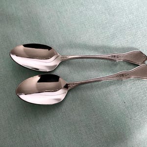 May include: Two shiny, silver spoons with a reflective surface are displayed on a light blue background. The spoons have a decorative floral design on the handle and a rounded bowl. The spoons are likely for serving or eating.