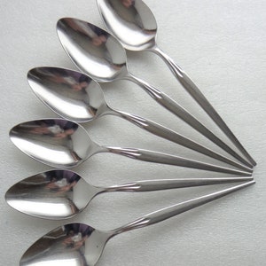 May include: A set of six shiny, stainless steel spoons arranged in a fan shape. Each spoon has a rounded bowl and a slender handle with a decorative ridge near the bowl. The spoons reflect the surrounding light.