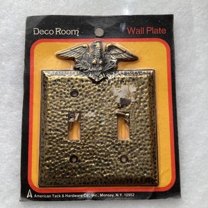May include: A brass-colored metal double toggle wall plate with a hammered texture and an eagle emblem at the top. The product is packaged on a black and yellow card labeled Deco Room Wall Plate by American Tack & Hardware Co., Inc., Monsey, N.Y. 10952.