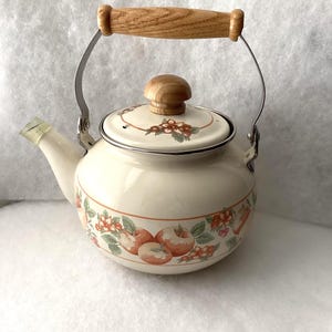 May include: A cream-colored enamel tea kettle featuring a decorative band of red apples and green leaves around the base. The kettle has a metal handle with a wooden grip, a matching wooden lid knob, and a small clear plastic spout cover.