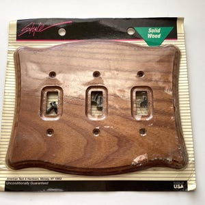 May include: A solid wood, triple switch plate with a dark brown finish. The plate has three rectangular openings for switches and screw holes. The packaging includes the text "Solid Wood" and "Made in USA".