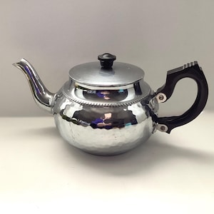 May include: A silver-colored teapot with a hammered texture. It has a black handle and lid knob. The spout is curved, and the lid sits atop the rounded body. The teapot is designed for brewing tea.