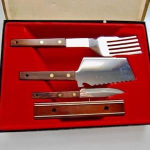 May include: A vintage barbecue tool set in a red velvet-lined box. The set includes a spatula, a knife, a pie server, and a fork. The tools have wooden handles with brass rivets and stainless steel blades.