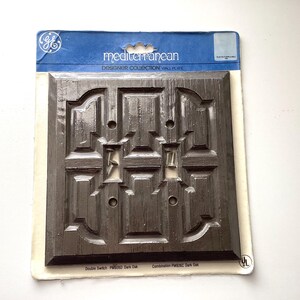 May include: A dark brown double switch wall plate from the Mediterranean Designer Collection. The plate has a detailed, carved design and is packaged on a blue and white card. The text "Mediterranean" is visible on the packaging.