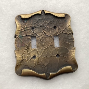 May include: A double light switch wall plate with a dark, aged brass finish and a textured, crinkled surface. The rectangular plate features decorative curved edges and four screw holes, with two vertical rectangular cutouts for switches.