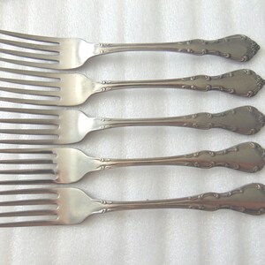 May include: Five silver forks with ornate handles are arranged on a white surface. The forks have multiple tines and decorative detailing on the handles. The silverware is likely for a formal dining setting.