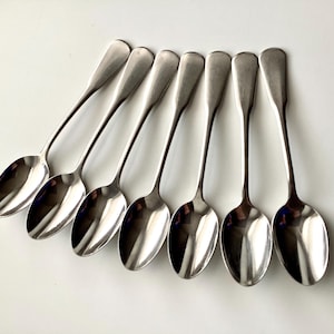 May include: Seven shiny, silver spoons arranged diagonally across a white surface. Each spoon has a rounded bowl and a long, slender handle. The spoons reflect light, creating highlights and shadows.