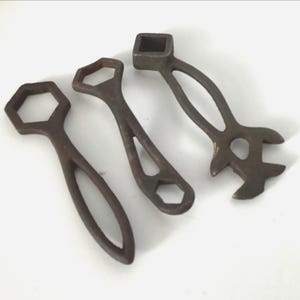 May include: Three antique, brown metal tools with open-ended designs. The tools include a hexagonal wrench, a wrench with a square opening, and a wrench with a curved handle. The tools are arranged on a white surface.