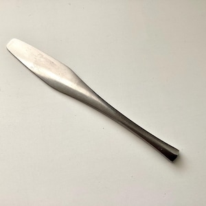 May include: A stainless steel butter knife with a rounded blade and a long, slender handle. The knife is designed for spreading and serving butter or other spreads. The utensil is made of a shiny, silver-colored metal.