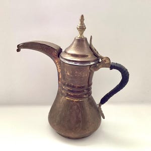 May include: Antique brass coffee pot with a long spout and a decorative lid. The pot features intricate etched patterns and a black wrapped handle. The design is reminiscent of traditional Middle Eastern coffee pots.