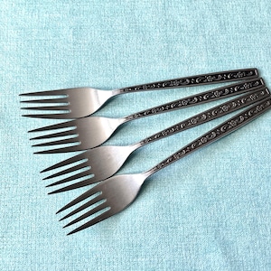 May include: Four stainless steel forks with a floral design on the handles. The forks are arranged diagonally on a light blue textured surface. The tines are long and slender.