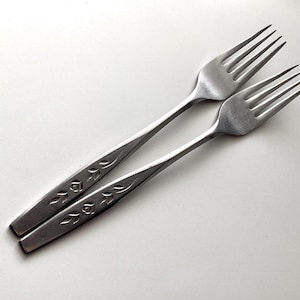 May include: Two stainless steel forks with four tines each, resting on a white surface. The handles feature an embossed floral design. The forks are angled towards the upper right, showcasing their polished finish and elegant design.