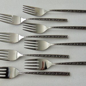 May include: A collection of eight stainless steel forks. Each fork has a three-tine design and a decorative floral pattern along the handle. The forks are arranged on a white surface.