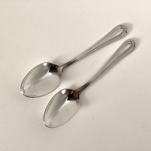 May include: Two stainless steel spoons with oval bowls and elongated handles. The handles have a decorative looped design at the end. The spoons are set against a white background, reflecting light.
