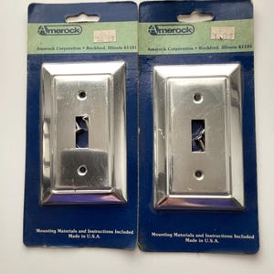 May include: Two silver-colored light switch covers on blue cardboard backing. The covers have a rectangular shape with a single switch opening. The packaging includes the Amerock logo and text that reads "Mounting Materials and Instructions Included Made in U.S.A."