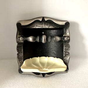 May include: A decorative wall-mounted soap dish with a silver-colored frame and a black interior. The dish features a cream-colored, shell-shaped soap holder and a silver-toned bar across the center. The frame has a textured, ornate design.