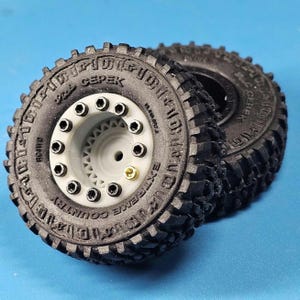 Puede incluir: Dos neumáticos todoterreno negros con el texto "CEPEK EXTREME COUNTRY" en los laterales. Las ruedas tienen un centro gris con pernos negros y una válvula dorada. Los neumáticos están diseñados para terrenos accidentados.