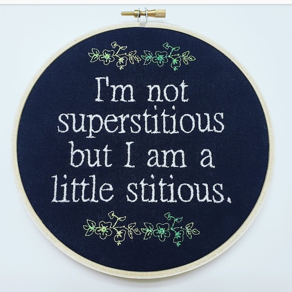 May include: Circular embroidery hoop featuring navy blue fabric with white text: "I'm not superstitious but I am a little stitious." Green and gold floral embroidery decorates the top and bottom. The hoop is made of wood.