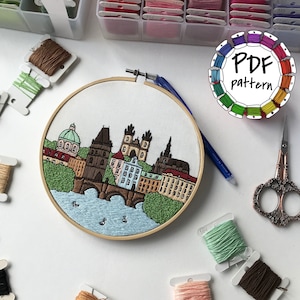 May include: Embroidered hoop art featuring a cityscape of Prague, Czech Republic. The scene includes the Charles Bridge, buildings, and a river. The hoop is surrounded by embroidery floss spools in various colors.