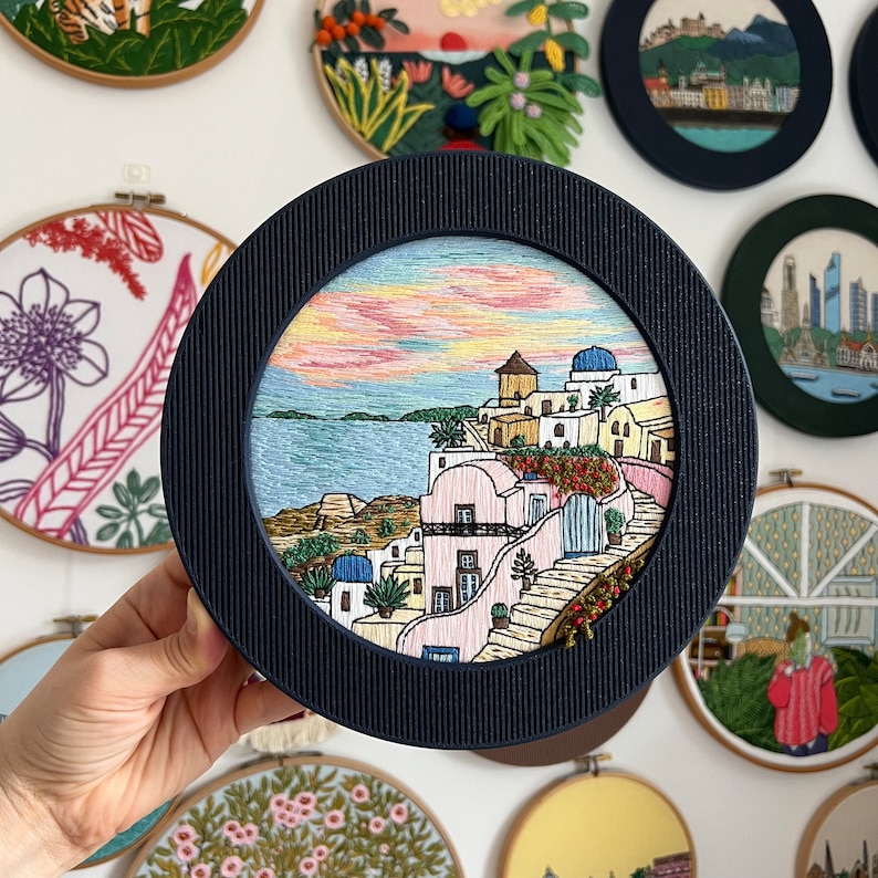 A hand holds a wooden display frame vertically, giving a clear view of its depth and smooth circular edge. Inside is an intricate embroidery of Santorini made with embroidery pattern by Fraimyxstitch. The background is a wall full of colorful embroideries. Designed to pair with embroidery hoops, display frame helps convert stitched designs into easy wall hangings without needing tools.