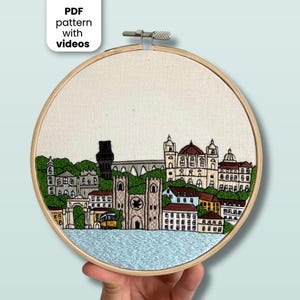 Op de afbeelding: Een ronde houten borduurring met een stoffen paneel waarop een handgeborduurde stadsgezicht te zien is. Het borduurwerk toont gebouwen, een brug en een gele tram in bruine, groene en witte tinten, boven blauw water. De tekst luidt: PDF pattern with videos.