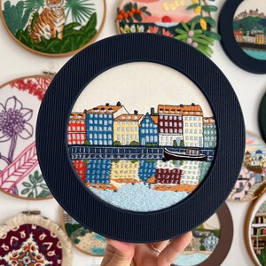 Puede incluir: Un aro de bordado azul marino con un fondo blanco que presenta una ciudad bordada en colores con edificios y un r&iacute;o. La ciudad est&aacute; rodeada por un borde blanco.